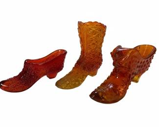 This collection features three decorative glass shoe and boot figures, likely intended as small vases or decorative accents. The items are crafted from pressed glass, exhibiting the characteristic Daisy and Button pattern, which consists of raised, star-like motifs and hobnail texture. The colors range from a deep amber to a lighter, more translucent orange-yellow, suggesting a gradient effect or variations in the glass mixture. The styles include a single shoe, a cowboy boot, and another ankle boot, each with a molded heel and intricate detailing in the pattern. These pieces are representative of Fenton Art Glass, popular for their decorative appeal and often produced in a variety of colors and forms.