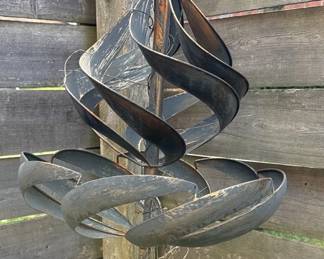 This is a metal kinetic wind sculpture, designed for garden or outdoor display. It features stylized tulip petal forms that rotate with the wind, along with stylized leaf forms. The material appears to be weathered steel or a similar metal, exhibiting a patina and some signs of rust, which is common for outdoor metal art exposed to the elements. The sculpture is mounted on a central pole, suggesting it is intended to be anchored in the ground.