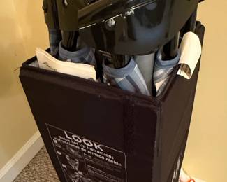 This is an Eddie Bauer Complete Care Play Yard, designed for versatile use as a playpen or crib. The item appears to be in a folded, stored state, with its black fabric cover and associated instruction manual visible. The play yard features a sturdy metal frame, with black plastic accents on the upper structure, and the fabric is a dark color with a plaid interior lining visible. The included manual, titled 'Complete Care Play Yard Quick Set Up Instructions', indicates it is a user guide for assembly and use, with a website listed for further assistance. The product is designed for safety and convenience, with warnings about proper assembly and use to prevent injury.