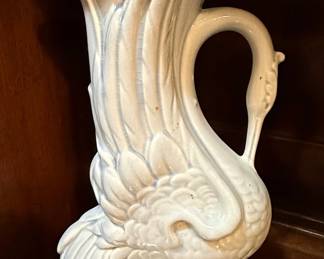 This vintage ceramic vase features a graceful swan design, with its body forming the base and its neck curving upwards to create the vase opening. The glaze appears to be a pale, almost iridescent white with subtle blue undertones, exhibiting a fine crackle pattern typical of older ceramic pieces. The detailing on the swan's feathers and head is rendered with a soft, artistic touch. The overall form suggests a decorative piece, likely intended for display rather than heavy use, with a classic aesthetic that could fit into various decor styles. Likely made by McCoy Pottery, a well-known American ceramic manufacturer.