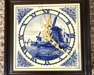 This is a decorative wall clock featuring a Delft-Style Blue ceramic tile face. The tile depicts a traditional Dutch landscape with windmills, water, and buildings, rendered in the characteristic blue and white palette. The clock is housed within a dark wooden frame. The clock face itself is ceramic, with Roman numerals marking the hours and ornate brass hands indicating the time. The style is reminiscent of traditional Dutch pottery, often referred to as Delftware.