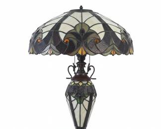 This table lamp features a dome-shaped shade crafted from numerous pieces of hand-cut stained glass in a floral and geometric pattern, with colors including cream, deep green, amber, and dark brown. A central amber jewel accentuates the base of the shade. The lamp's base and stem are made of dark, patinated metal with ornate scrollwork and a tiered foot. This lamp is likely the Chloe Liaison or River of Goods Halston model.