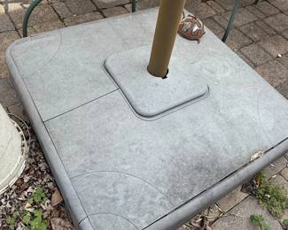This is a cantilever or offset patio umbrella, characterized by its side-mounted pole that allows the canopy to extend over a seating or dining area without a central pole obstruction. The umbrella features a large, cream-colored fabric canopy, currently closed and tied. The supporting structure appears to be made of dark metal, likely aluminum or steel, with a rust-resistant coating. The base is a substantial, multi-piece cast iron construction designed to provide stability for the large umbrella. The individual base segments are decorative, featuring a fan-like or sunburst pattern. The overall style suggests a functional and decorative outdoor furnishing suitable for patios, decks, or gardens.