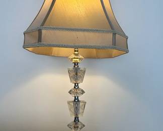 This vintage table lamp features a striking design composed of stacked cut crystal elements, culminating in a faceted base and a decorative finial. The central stem is adorned with geometric crystal prisms, each meticulously cut to refract light. A prominent feature is the etched geometric pattern on one of the crystal sections, adding a layer of intricate detail and visual interest. The lamp is topped with a classic bell-shaped lampshade in a neutral tone, trimmed with decorative braiding. The overall aesthetic suggests a mid-20th-century origin, likely influenced by Art Deco or Hollywood Regency styles, emphasizing glamour and sophisticated ornamentation.