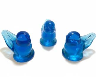 A set of three small, translucent blue glass figurines, each depicting a stylized bluebird. The birds have a smooth, rounded body, folded wings, and a simple, uplifted tail. They are designed to stand upright on a flat base. The glass has a vibrant cobalt blue hue, allowing light to pass through and creating a luminous effect. One of the figurines still retains its original "Sunny Day Bluebirdâ„¢" sticker, featuring a cheerful sun and bird graphic, which helps in identifying the maker and product line. These decorative pieces are commonly used as collectibles or as accents for home decor. The "Sunny Day Bluebirdâ„¢" brand is associated with the "Bluebird of Happiness" collectible series, often created by artists like Leo Ward and Ron Ray.