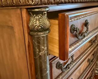 This bedroom set, comprising a tall chest of drawers, a nightstand, and a dresser with a mirror, is crafted from cherry wood with a rich, warm finish. The style is reminiscent of Italian Baroque, characterized by ornate carvings, scrolled hardware, and decorative inlays. Each piece features faux marble columns with detailed capitals and bases, adding a touch of opulence. The dresser top and nightstand top appear to be made of real marble, likely black with white veining, providing a durable and luxurious surface. The intricate detailing on the drawer fronts and the central decorative panel of the dresser suggests a high level of craftsmanship, typical of furniture designed to make a statement.