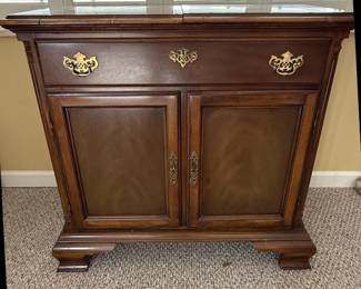 This American Drew sideboard, part of the "American Independence Collection," is crafted from rich, dark wood, likely mahogany or a high-quality veneer, showcasing a traditional style. It features a central drawer with ornate brass hardware, flanked by two cabinet doors with inset panels that exhibit subtle wood grain patterns. The base has a decorative, stepped plinth, and the overall construction suggests a durable and elegant piece of furniture. The "American Independence Collection" line is inspired by the original furnishings of Independence Hall, aiming to capture a sense of historical significance and classic American design. The brass hardware, with its intricate scrollwork, complements the dark wood finish, creating a piece that is both functional and decorative.