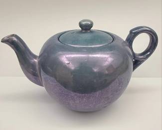 This vintage teapot features a striking lusterware glaze that shifts between shades of purple and teal. The glaze is applied unevenly, with a more concentrated purple hue towards the base, transitioning to a cooler teal on the upper body and lid. The teapot has a classic, rounded body with a gracefully curved spout and a C-shaped handle, typical of many ceramic teapots produced in the mid-20th century. The underside bears a "Made in Japan" mark. The overall aesthetic suggests a decorative piece, possibly from the Mid-Century Modern era, designed to be as visually appealing as it is functional.