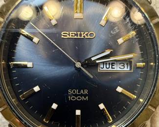 This lot features two men's Seiko wristwatches. The first is a solar-powered watch with a dark blue dial, gold-tone hour markers and hands, and a day-date complication. It indicates 'SOLAR 100M' on the dial and 'MOV'T JAPAN V158-0AD0 R 2' on the case back, which is stainless steel. The second watch has a white dial with gold-tone hour markers and hands, and a day-date complication, and 'WATER RESIST 50M' printed on the dial. Its case back indicates 'MOV'T JAPAN V742-2161 R 2'. Both watches have gold-tone metal bracelets and cases. The overall aesthetic suggests a classic, functional design typical of Seiko's offerings in the late 20th or early 21st century. The crystals on both watches show significant scratching.
