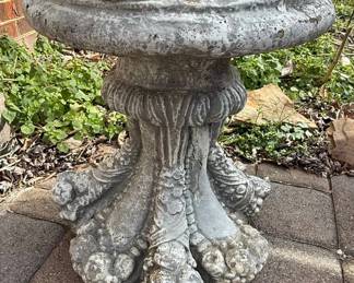 This cast stone side table features a substantial, circular basin with a slightly beveled edge, supported by an intricately sculpted pedestal. The stem exhibits fluted detailing, transitioning into a robust lower section adorned with four prominent claw feet, each appearing to grip a decorative orb or cluster. The material is weathered, showing signs of age and exposure, with a patina that highlights its texture and form. This piece evokes a classical or gothic aesthetic, commonly used as a plinth for statues, urns, or as a decorative garden accent.