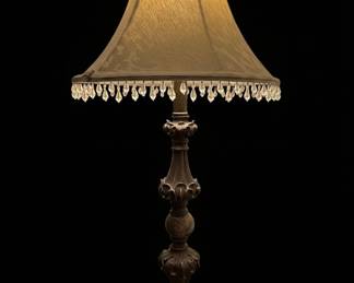 This vintage table lamp features a classic bell-shaped shade crafted from a textured, light gold fabric, adorned with a delicate fringe of clear crystal beads. The base and stem are made of a dark, patinated metal, likely bronze or brass, with an ornate, carved design reminiscent of acanthus leaves and scrollwork. The overall aesthetic suggests a Victorian or Edwardian influence, designed to add a touch of traditional elegance to a room. The crystal beads catch and refract light, creating a subtle sparkle. The lamp appears to be in good condition, though the fabric shade may show signs of age and dust. The ornate base with acanthus leaf carvings is a prominent feature.