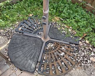This is a cantilever or offset patio umbrella, characterized by its side-mounted pole that allows the canopy to extend over a seating or dining area without a central pole obstruction. The umbrella features a large, cream-colored fabric canopy, currently closed and tied. The supporting structure appears to be made of dark metal, likely aluminum or steel, with a rust-resistant coating. The base is a substantial, multi-piece cast iron construction designed to provide stability for the large umbrella. The individual base segments are decorative, featuring a fan-like or sunburst pattern. The overall style suggests a functional and decorative outdoor furnishing suitable for patios, decks, or gardens.