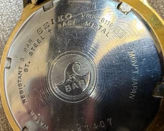 This lot features two men's Seiko wristwatches. The first is a solar-powered watch with a dark blue dial, gold-tone hour markers and hands, and a day-date complication. It indicates 'SOLAR 100M' on the dial and 'MOV'T JAPAN V158-0AD0 R 2' on the case back, which is stainless steel. The second watch has a white dial with gold-tone hour markers and hands, and a day-date complication, and 'WATER RESIST 50M' printed on the dial. Its case back indicates 'MOV'T JAPAN V742-2161 R 2'. Both watches have gold-tone metal bracelets and cases. The overall aesthetic suggests a classic, functional design typical of Seiko's offerings in the late 20th or early 21st century. The crystals on both watches show significant scratching.