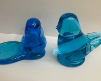 This set features two charming glass figurines, a "Bluebird of Happiness" and a companion piece, both crafted from vibrant blue glass. The larger "Bluebird of Happiness" is a classic representation, with its stylized wings and tail. The smaller piece appears to be a nest or a base, designed to complement the bluebird. Both pieces are made of solid, translucent blue glass, exhibiting a smooth finish with some internal bubbles characteristic of hand-blown glass. The "Bluebird of Happiness" is known for its uplifting symbolism and has been a popular decorative item for decades, often associated with good fortune and joy. The set is signed by Terra Studio and includes original labeling.
