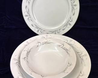 This set includes a lidded sugar bowl, a creamer pitcher, and a teacup with its matching saucer, all crafted from fine china. The pattern, "Platinum Garland," features delicate platinum trim along the edges and a graceful design of wheat stalks and leaves in a muted gray and subtle copper hue. The pieces are white with a smooth, glossy finish, indicative of quality porcelain. The sugar bowl has elegant, curved handles and a decorative lid finial. The creamer exhibits a classic spout and a single handle, while the teacup has a matching handle and sits on a saucer adorned with the same garland motif. The maker's mark on the underside of the teacup identifies it as Harmony House Fine China, Platinum Garland, Made in Japan, with the pattern number 3541.