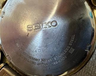 This lot features two men's Seiko wristwatches. The first is a solar-powered watch with a dark blue dial, gold-tone hour markers and hands, and a day-date complication. It indicates 'SOLAR 100M' on the dial and 'MOV'T JAPAN V158-0AD0 R 2' on the case back, which is stainless steel. The second watch has a white dial with gold-tone hour markers and hands, and a day-date complication, and 'WATER RESIST 50M' printed on the dial. Its case back indicates 'MOV'T JAPAN V742-2161 R 2'. Both watches have gold-tone metal bracelets and cases. The overall aesthetic suggests a classic, functional design typical of Seiko's offerings in the late 20th or early 21st century. The crystals on both watches show significant scratching.