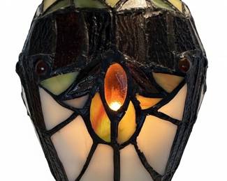 This table lamp features a dome-shaped shade crafted from numerous pieces of hand-cut stained glass in a floral and geometric pattern, with colors including cream, deep green, amber, and dark brown. A central amber jewel accentuates the base of the shade. The lamp's base and stem are made of dark, patinated metal with ornate scrollwork and a tiered foot. This lamp is likely the Chloe Liaison or River of Goods Halston model.