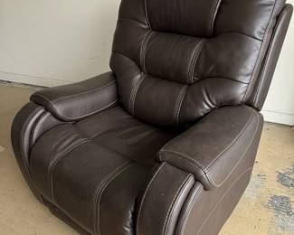 This is a brown, faux leather power lift recliner chair. It features plush, overstuffed cushioning with decorative stitching in a contrasting cream color. The chair is operated by a wired remote control that allows for adjustment of the headrest and back, as well as a lift function, indicated by directional arrows on the remote. The remote also has buttons for headrest and back adjustments, suggesting multiple reclining positions and lumbar support options. The material appears to be a durable, easy-to-clean faux leather, suitable for everyday use. The overall design is contemporary and emphasizes comfort and ease of use, particularly for individuals who may require assistance with standing or sitting.
