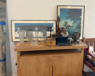 Wooden hand carved yoga meditation figurine, brass or bronze bell, candlestick holder, a knotty old man handmade collector candle, Balinese cast iron door stopper, Maxfield Parrish twilight art, white Canadian barn by Georgia O’Keeffe, mid-century Oak Brown wooden credenza, crystal vases.