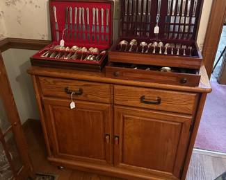 Oak server cabinet on wheels displaying two silverware sets.