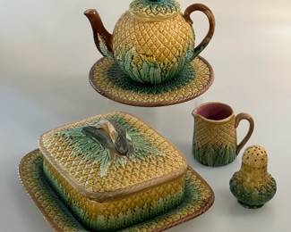 Antique majolica pineapple pattern items circa 1880 in green, yellow and brown comprising a lidded teapot and tea tile, a covered pineapple pattern box with attached underplate and lid with with gray colored sardine handles, a creamer standing 2-¼ inches H, and a 3-inch tall pepper pot.