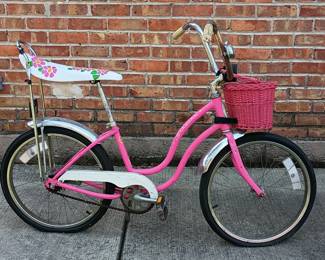 A 1960s vintage pink Schwinn Sting-Ray bicycle with a distinctive "banana" seat with a floral pattern and a high backrest; equipped with a pink wicker-style front basket and chrome fenders.