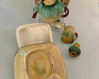 Reverse view of antique majolica pineapple pattern items circa 1880 in green, yellow and brown comprising a lidded teapot and tea tile, a covered pineapple pattern box with attached underplate and lid with with gray colored sardine handles, a creamer standing 2-¼ inches H, and a 3-inch tall pepper pot.