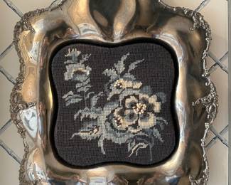 A silver plate tray with fruiting grape vine on the scalloped edges and a floral design needlepoint insert