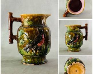 A large majolica earthenware pitcher with raised figure of a bird resting on leafy branches, richly glazed/hand painted in mottled/streaking background of shades of green and brown; bird has black wings and head with deep rose breast; brown branch handle and lavender liner. 9-1/4 inch high.
