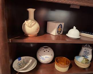 TOP SHELF /Red Wing Pottery Magnolia line vase in ivory $60.00 |  Fleur-de-lis Square Planter: A heavy white ceramic planter featuring a raised gold fleur-de-lis emblem set within a deep blue oval medallion. Price: $45.00 | Bird-Finial Porcelain Dome: A dainty white porcelain lidded dish topped with a sculpted bird figurine. Price: $30.00 | Geometric Blue-Rimmed Tray: A slender, rectangular porcelain tray accented with a blue-and-white patterned border. Price: $25.00 |  BOTTOM SHELF / Crane Motif Perforated Globular Vase: A round white porcelain vessel decorated with blue flying cranes and circular perforations for incense or floral stems. Price: $45.00 |   luted Brown Glaze Ramekin: A sturdy ceramic baking dish with a cream-colored fluted exterior and a rich brown glazed interior. Price: $28.00 | lue Floral "Caffe" Jar: A vintage-style porcelain jar featuring blue floral flourishes $25.00 | 2 Small ceramic bowls with interior painting set $27.00 