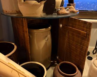 Circular Glass Display Top: A thick, tempered clear glass round sheet Price $120 | Wooden Crate Base: A weathered wooden shipping crate used as a rustic pedestal,$75.00 | Tan 6-gallon ceramic Vessel housed into the crate $495 | Two-Tone Handled Glazed Pot: A wide-mouthed ceramic vessel with a deep chocolate-brown glazed rim and handles $55 (on the table) | Miniature Wooden Shoe: A pair of small, hand-carved vintage wooden shoe molds with weathered patina Price: $50.00 (Pair) | (on the floor) Antique Brown ceramic unglazed Pot $125