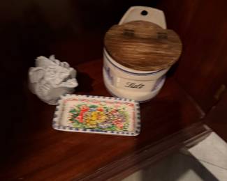 White Ceramic Gargoyle Bust: A small, detailed gothic-style gargoyle sculpture with an unglazed, matte finish. Price: $20.00 | Hand-Painted Floral Rectangular Dish: A decorative ceramic tray featuring vibrant, multi-colored floral artwork and a blue-and-white dotted border. Price: $22.00 | Vintage Ceramic Salt Cellar: A white stoneware crock labeled "Salt" in blue script, featuring a hinged wooden lid and a pre-drilled hole for wall mounting. Price: $35.00  