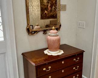 A pink ceramic ginger jar sits atop Councill Craftsmen four-drawer bachelor chest all below a gold gilt Rococo-style mirror.