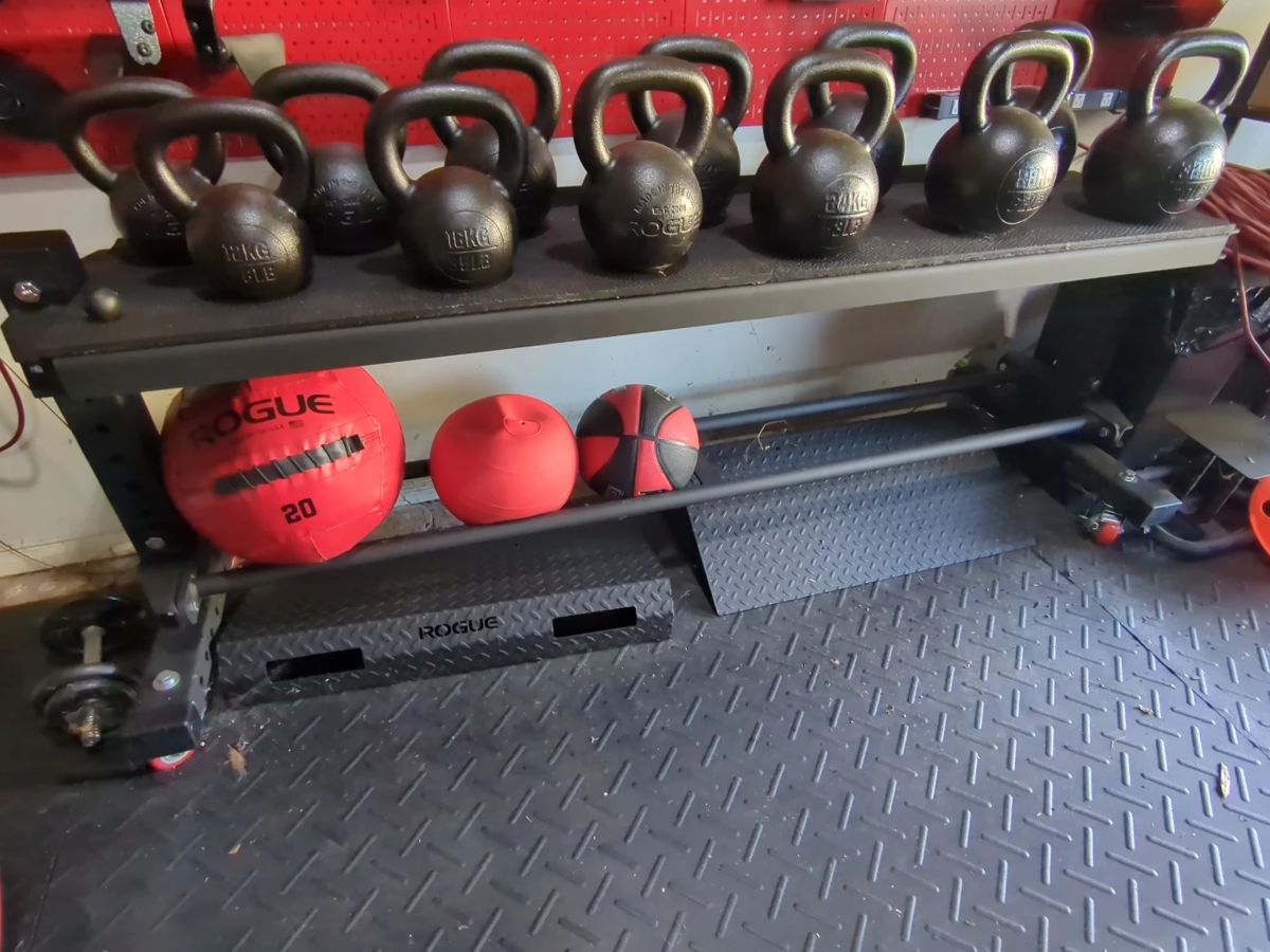 Lot 43:Set of Rogue kettle weights on rolling stand ranging from 25 lb to 70 lb. Includes pair of rogue diamond plate, metal ramps underneath and balls. All for your bid