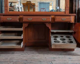 A large mahogany sideboard with an attached mirror by Aldis & Leggott, cabinet makers from Hull. The piece features four drawers, two side cabinets with pull-out shelves, and a specialized interior section for bottle storage. Decorative carvings on the legs and facade, as well as brass drawer pulls, highlight the craftsmanship. Dovetail joinery is observed on the drawers, indicating quality construction. The mirror shows minor silvering loss, and the wooden structure displays some signs of wear consistent with age, including small scratches and woodworm holes. Stamped “Aldis & Leggott, 17 Paragon Street, Hull”. This stunning sideboard measures 77” H X 78” W x 26” D.