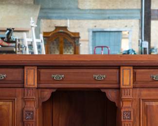 A large mahogany sideboard with an attached mirror by Aldis & Leggott, cabinet makers from Hull. The piece features four drawers, two side cabinets with pull-out shelves, and a specialized interior section for bottle storage. Decorative carvings on the legs and facade, as well as brass drawer pulls, highlight the craftsmanship. Dovetail joinery is observed on the drawers, indicating quality construction. The mirror shows minor silvering loss, and the wooden structure displays some signs of wear consistent with age, including small scratches and woodworm holes. Stamped “Aldis & Leggott, 17 Paragon Street, Hull”. This stunning sideboard measures 77” H X 78” W x 26” D.