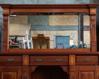 A large mahogany sideboard with an attached mirror by Aldis & Leggott, cabinet makers from Hull. The piece features four drawers, two side cabinets with pull-out shelves, and a specialized interior section for bottle storage. Decorative carvings on the legs and facade, as well as brass drawer pulls, highlight the craftsmanship. Dovetail joinery is observed on the drawers, indicating quality construction. The mirror shows minor silvering loss, and the wooden structure displays some signs of wear consistent with age, including small scratches and woodworm holes. Stamped “Aldis & Leggott, 17 Paragon Street, Hull”. This stunning sideboard measures 77” H X 78” W x 26” D.