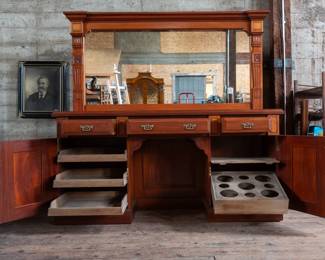A large mahogany sideboard with an attached mirror by Aldis & Leggott, cabinet makers from Hull. The piece features four drawers, two side cabinets with pull-out shelves, and a specialized interior section for bottle storage. Decorative carvings on the legs and facade, as well as brass drawer pulls, highlight the craftsmanship. Dovetail joinery is observed on the drawers, indicating quality construction. The mirror shows minor silvering loss, and the wooden structure displays some signs of wear consistent with age, including small scratches and woodworm holes. Stamped “Aldis & Leggott, 17 Paragon Street, Hull”. This stunning sideboard measures 77” H X 78” W x 26” D.