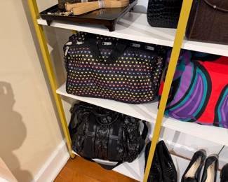 Black LeSportsac duffel bag with multicolored heart print sits beside a bold red-purple graphic tote, with a crinkled black drawstring bag stored below.