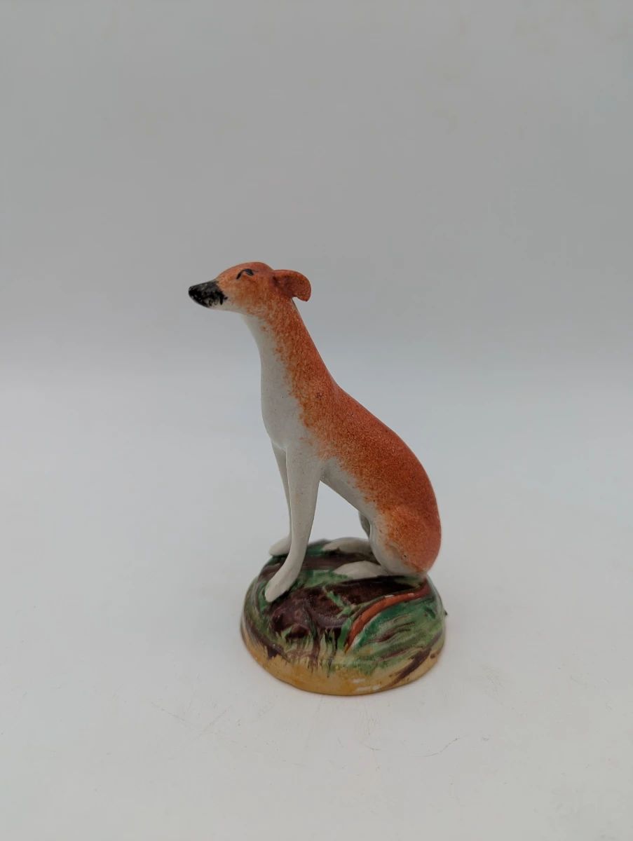 Small Victorian Staffordshire pottery greyhound figure, circa late 19th century. Hand-painted in orange and white with green and brown mound base. Measures 4 inches tall.