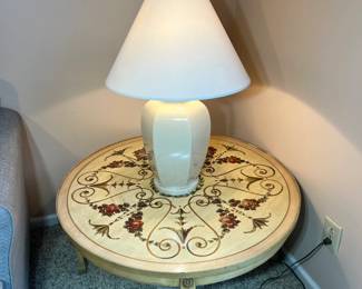 A round, cream-painted end table featuring an intricate hand-painted floral and scrollwork design and lamp with shade