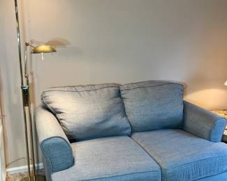 A tall, sleek brass floor lamp with an adjustable "pharmacy style" arm, shown next to a plush gray loveseat