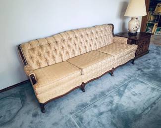 Stunning vintage Hollywood Regency / Mid-Century Modern tufted sofa. Features an elegant curved silhouette upholstered in a rich, shimmering gold satin fabric. Fully designed with deep button tufting across the backrest, a comfortable three-cushion seat configuration. Supported by a beautifully carved dark wood accent trim along the front armrests and bottom edge. A high-end vintage statement seating piece in exceptional condition, perfect for adding classic retro luxury to any living space.
