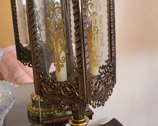 An ornate antique‑style lantern lamp with elaborate metalwork in a gold‑bronze finish. The frame features scrolling floral patterns, embossed detailing, and decorative glass panels with frosted or etched designs. Inside sits a candle‑shaped light fixture that adds to its Victorian or Old‑World character. The rounded, heavily detailed base gives it a substantial, high‑quality presence, making it ideal as a statement accent on a sideboard, entry table, or mantel.  Priced at $100