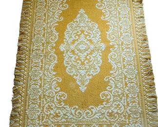 A rectangular decorative golden rug with an ornate white pattern featuring scrolling motifs, floral elements, and a central medallion design. The border repeats the same intricate detailing, and the edges are finished with a thick golden fringe. The overall look is traditional and elegant, making it suitable for a living room, bedroom, or as an accent under a small table or bench. The condition appears gently used, with the pattern still clear and the fringe intact.  Priced at