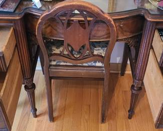 A vintage ornate wooden vanity desk with matching chair, featuring carved details, a curved front, and a glass‑top surface that protects the wood while showcasing its design. The vanity has two sets of drawers, all with decorative handles; the interiors are clean and functional, with one drawer currently holding small household items. The matching chair has an intricately carved backrest and a cushioned seat upholstered in a floral fabric, adding a traditional, elegant touch. The overall style leans toward French Provincial or classic vintage bedroom furniture, making it a charming statement piece for a bedroom, dressing area, or guest room.  Priced at $250