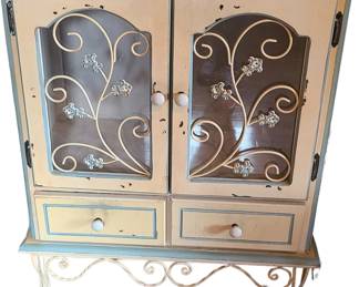 A vintage distressed cream and blue display cabinet with ornate detailing and a charming rustic‑elegant style. The two upper doors feature glass panels with decorative metal scrollwork and floral motifs, giving the piece a romantic, French‑inspired look. Below the doors are two storage drawers, and the entire cabinet stands on curved metal legs that echo the scroll design. The cream paint has a gently worn, intentionally aged finish with soft blue trim, adding character and making it a standout accent piece for cottage, shabby chic, or eclectic interiors.  Price at $75