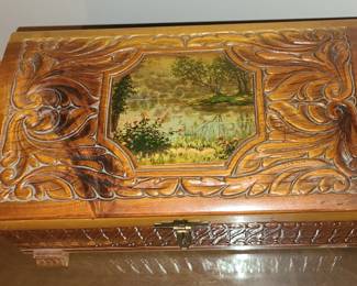 A carved wooden keepsake box featuring ornate swirling leaf patterns framing a hand‑painted landscape scene on the lid. The painting shows a peaceful natural setting with water, trees, and flowering foliage, giving the box a serene, decorative quality. The craftsmanship suggests a handcrafted piece, with detailed carving, a front latch closure, and a warm wood tone that adds to its vintage charm. It works well as a jewelry box, memory box, or decorative accent on a dresser or shelf.  Priced at $30