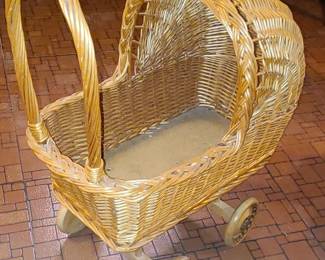 A small vintage wicker doll carriage made from woven rattan with a curved hood, wooden handle, and four wooden wheels. Its handcrafted look and natural patina give it charming cottage‑style appeal. This type of piece is typically decorative—used for dolls, display, or as a whimsical accent in a nursery, craft room, or vintage‑themed space.  Priced at $70
