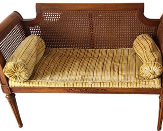 A vintage wooden cane‑back settee with elegant carved details and classic styling. The frame features woven cane panels along the back and sides, paired with carved accents on the front apron and tapered legs. The seat includes a striped cushion in soft yellow and beige tones, along with two matching bolster pillows that complete the traditional look. The piece has a light, airy feel thanks to the cane work, making it suitable for an entryway, sitting room, sunroom, or bedroom.